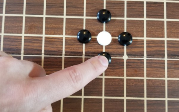 A hand points to one white stone, with 4 black stones surrounding it. this is called a liberty in the game of Go.